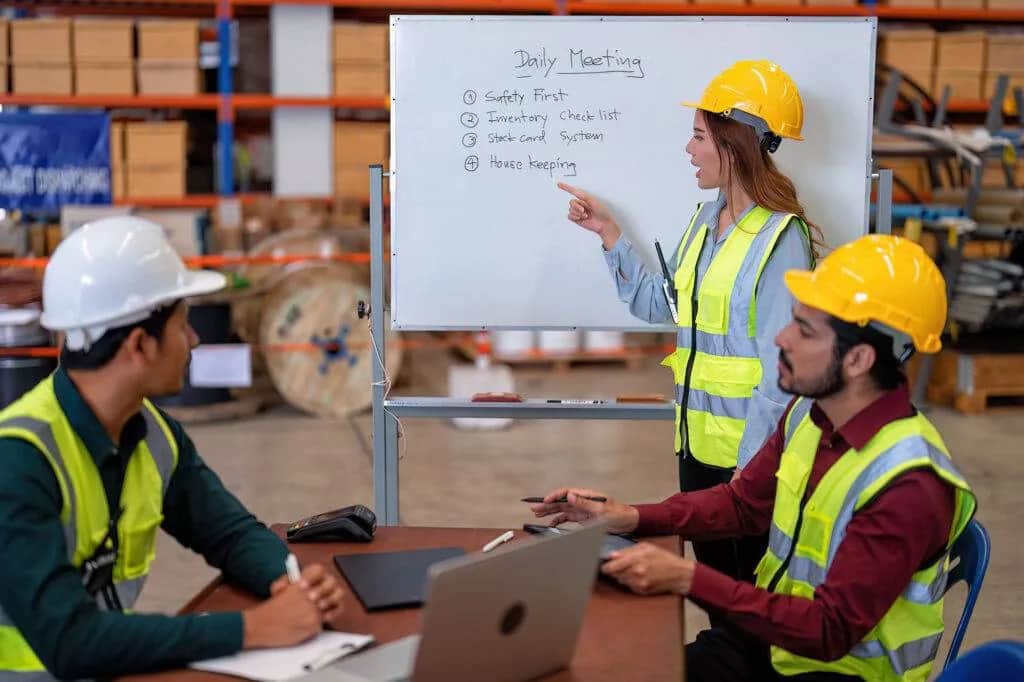 Supervisor in a hard hat and hi-vis vest leading a safety meeting at a whiteboard in a warehouse while two colleagues take notes
