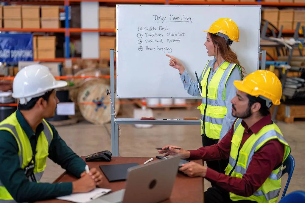 Supervisor in a hard hat and hi-vis vest leading a safety meeting at a whiteboard in a warehouse while two colleagues take notes