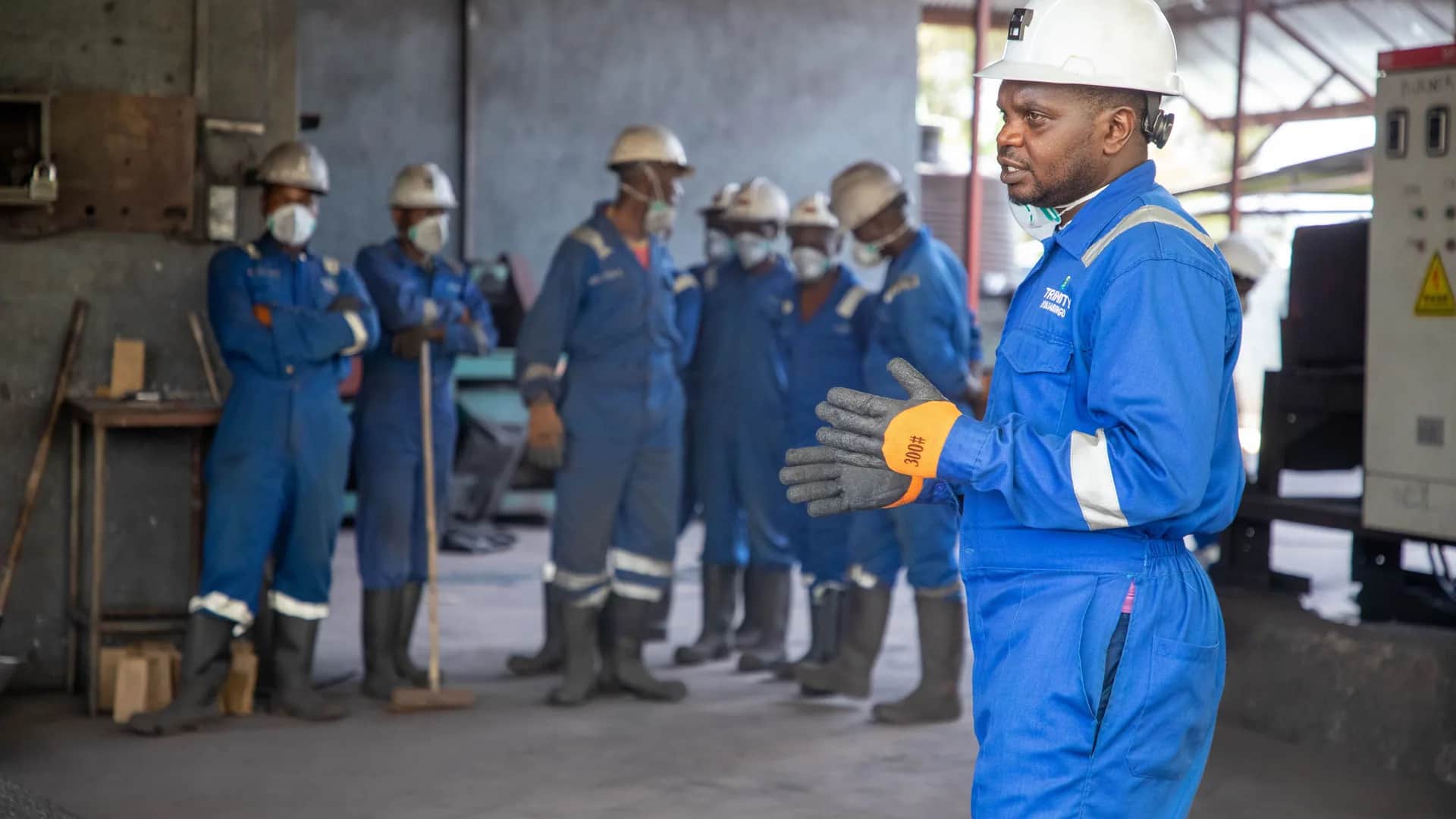 Trainer in a hard hat and gloves briefing a crew in coveralls, helmets and dust masks inside an industrial plant