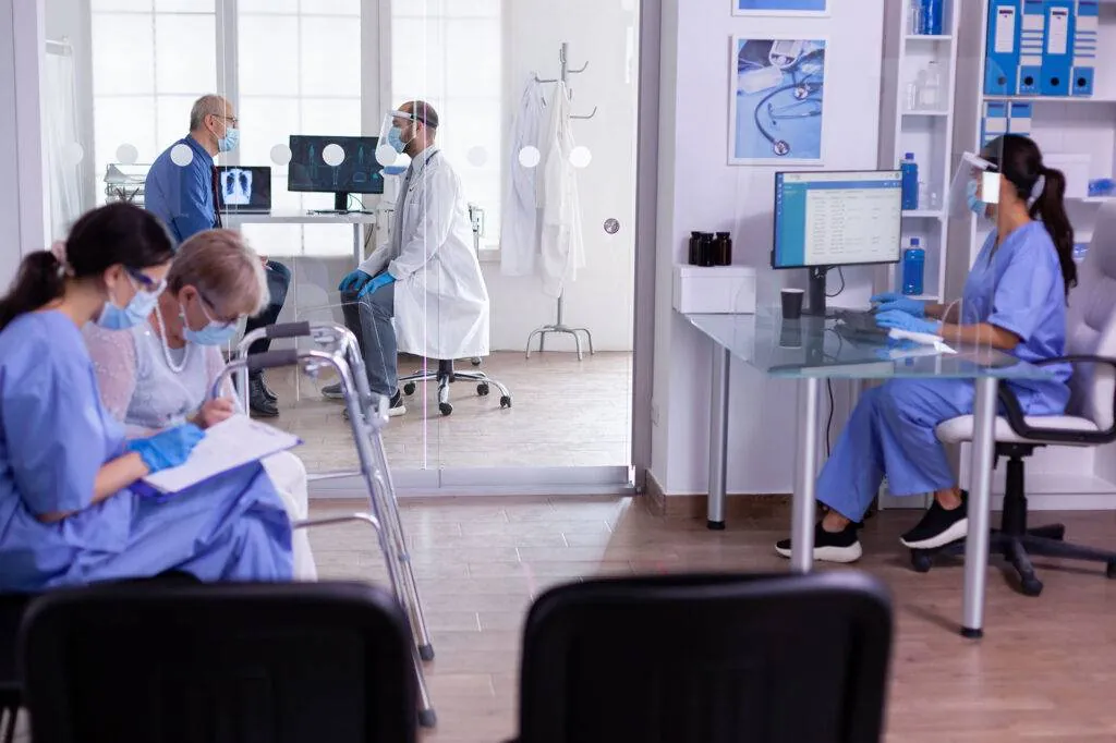 Clinic staff in scrubs, masks and face shields assisting patients in a waiting area and consultation room