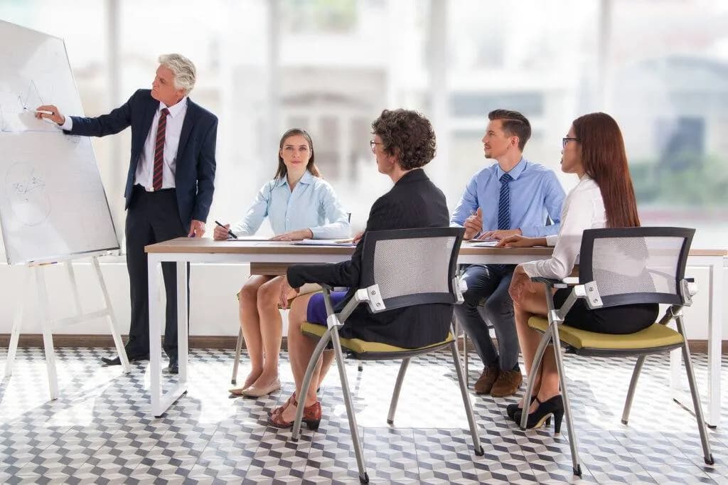 Trainer pointing at a flip chart while four delegates listen at a conference table in a bright training room
