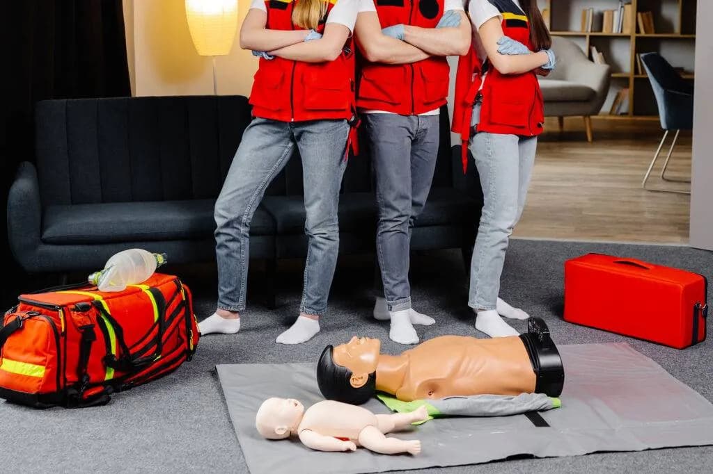 Adult and infant CPR manikins on a mat beside first aid bags as trainees in red vests and gloves stand by