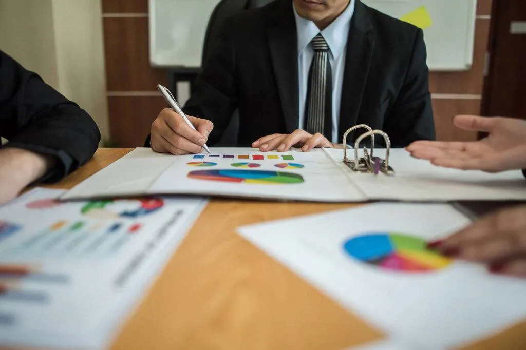 Professionals reviewing printed charts in a ring binder at a meeting table, one making notes with a pen