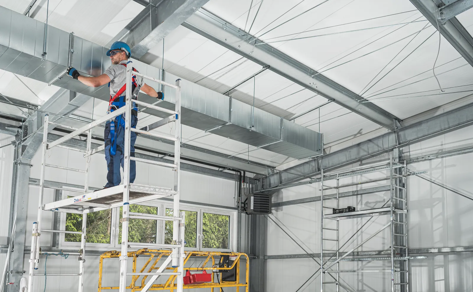 Worker in a fall-arrest harness standing on a mobile aluminium tower to fit ductwork in a steel-framed building
