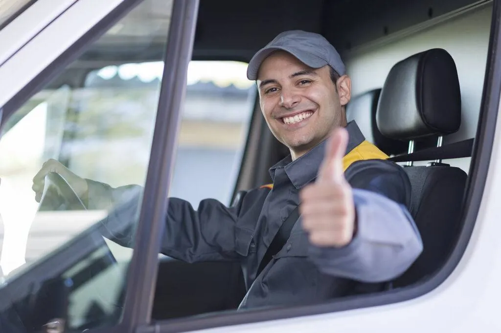 Smiling driver in a cap and work uniform giving a thumbs up from behind the wheel of a white van