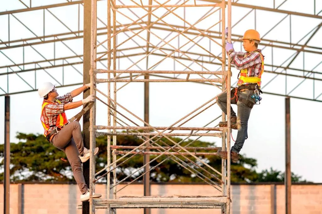 Two workers in hard hats and fall-arrest harnesses climbing a scaffold tower beneath a steel roof frame